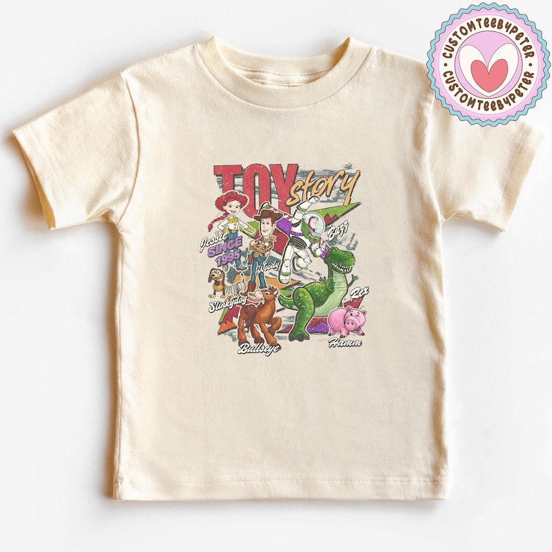 May include: A cream-colored short-sleeve t-shirt featuring a colorful graphic print of Toy Story characters including Woody, Buzz Lightyear, Jessie, Rex, Hamm, Bullseye, and Slinky Dog with the text Toy Story and individual character names.