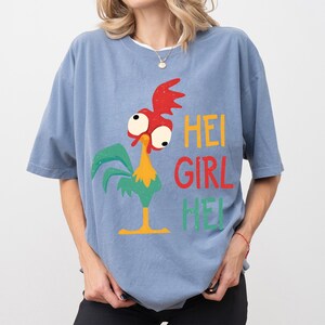 Peut inclure: T-shirt bleu clair avec un dessin de coq de dessin anim&eacute; et le texte "HEI GIRL HEI" en jaune, rouge et vert. Le coq a une cr&ecirc;te rouge, de grands yeux et des plumes vertes. Le t-shirt est de style d&eacute;contract&eacute;.
