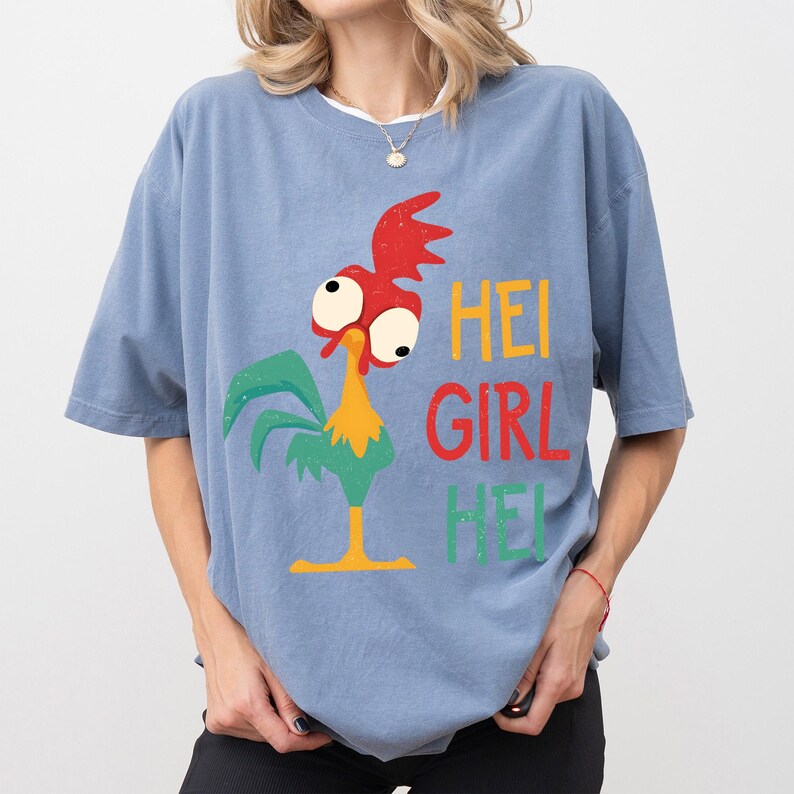 Peut inclure: T-shirt bleu clair avec un dessin de coq de dessin anim&eacute; et le texte "HEI GIRL HEI" en jaune, rouge et vert. Le coq a une cr&ecirc;te rouge, de grands yeux et des plumes vertes. Le t-shirt est de style d&eacute;contract&eacute;.