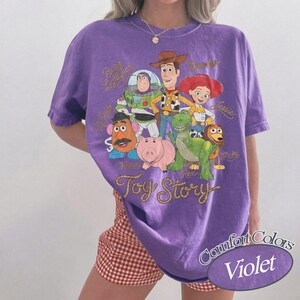 May include: A violet short-sleeve t-shirt featuring a graphic print of Toy Story characters including Buzz Lightyear, Woody, Jessie, Mr. Potato Head, Hamm, Rex, and Slinky Dog. The text Toy Story is printed in a gold cursive font below the characters.