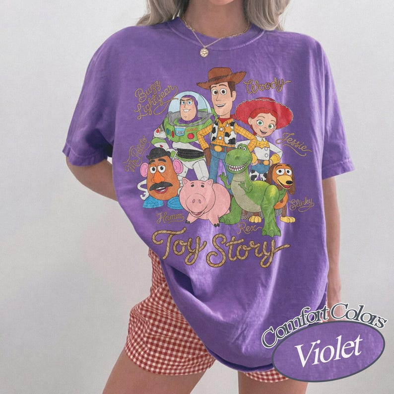 May include: A violet short-sleeve t-shirt featuring a graphic print of Toy Story characters including Buzz Lightyear, Woody, Jessie, Mr. Potato Head, Hamm, Rex, and Slinky Dog. The text Toy Story is printed in a gold cursive font below the characters.