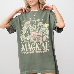 May include: Khaki green t-shirt with a colourful graphic design featuring a castle, Mickey Mouse, and the text "The Most MAGICAL PLACE". The design includes pink, yellow, and white accents. The shirt has short sleeves and a crew neck.