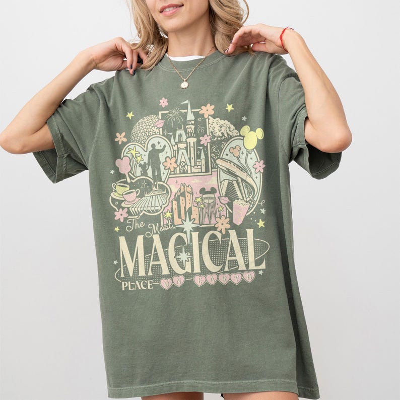 May include: Khaki green t-shirt with a colourful graphic design featuring a castle, Mickey Mouse, and the text "The Most MAGICAL PLACE". The design includes pink, yellow, and white accents. The shirt has short sleeves and a crew neck.