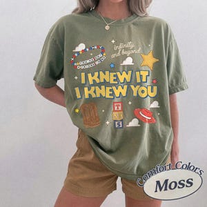 May include: An olive green t-shirt featuring a graphic print with the text "I KNEW IT I KNEW YOU", "infinity and beyond", and "YOU'VE GOT A FRIEND IN ME". The design includes illustrations of a beaded bracelet, a star, cowboy boots, blocks, and a hat.
