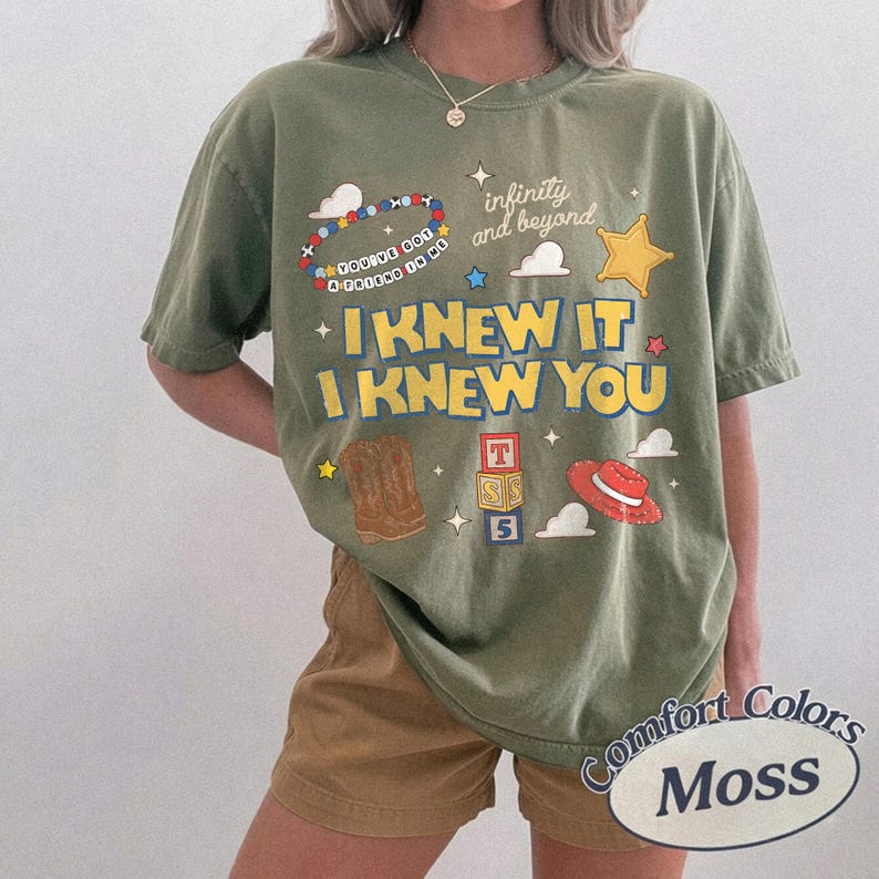 May include: An olive green t-shirt featuring a graphic print with the text "I KNEW IT I KNEW YOU", "infinity and beyond", and "YOU'VE GOT A FRIEND IN ME". The design includes illustrations of a beaded bracelet, a star, cowboy boots, blocks, and a hat.