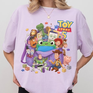 May include: A light purple t-shirt featuring a graphic print of various Toy Story characters including Woody, Buzz Lightyear, and a green frog emerging from a purple backpack, with the text Toy Story 5 printed in blue and yellow above the characters.