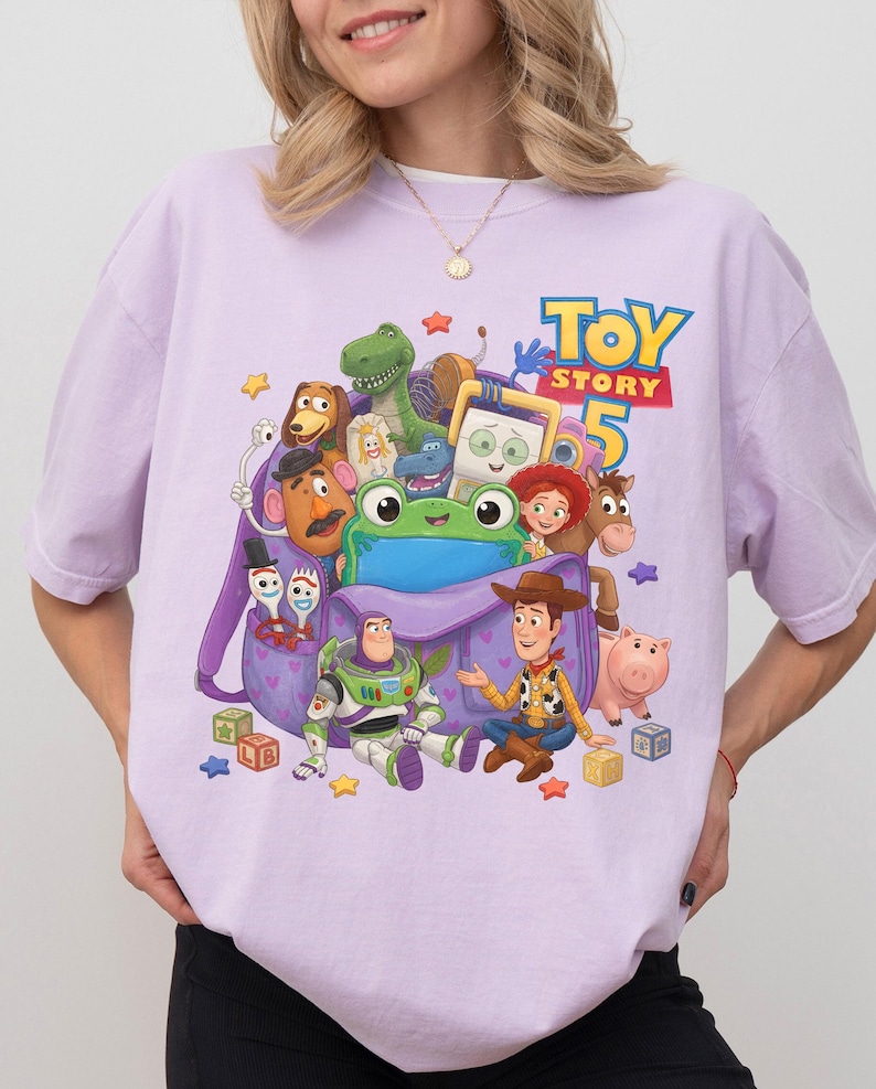 May include: A light purple t-shirt featuring a graphic print of various Toy Story characters including Woody, Buzz Lightyear, and a green frog emerging from a purple backpack, with the text Toy Story 5 printed in blue and yellow above the characters.