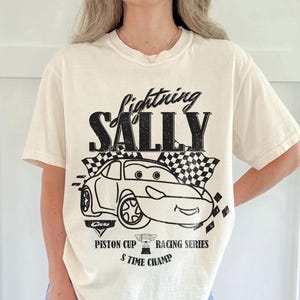 May include: Cream-coloured t-shirt with a black graphic of a cartoon car and the text "Lightning Sally." The design includes checkered flags and the words "Piston Cup Racing Series" and "5 Time Champ."
