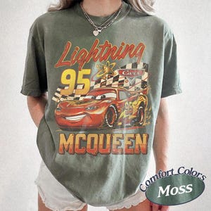 May include: A moss green t-shirt featuring a graphic of Lightning McQueen from the Cars movie. The design includes the text "Lightning 95 McQueen" in a vintage style, with a checkered flag and a racing car.