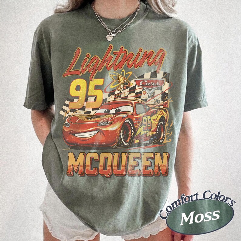 May include: A moss green t-shirt featuring a graphic of Lightning McQueen from the Cars movie. The design includes the text "Lightning 95 McQueen" in a vintage style, with a checkered flag and a racing car.