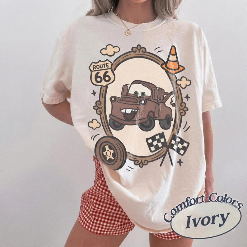 May include: Ivory-colored t-shirt featuring a cartoon image of a brown truck inside a decorative frame, with Route 66 signage, a traffic cone, and checkered flags. The shirt is labeled "Comfort Colors Ivory".