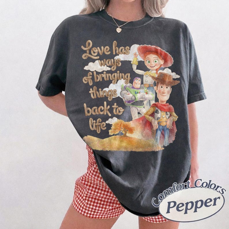 K&ouml;nnte beinhalten: Ein anthrazitfarbenes Kurzarm-T-Shirt mit einem Grafikdruck von Woody, Buzz Lightyear und Jessie aus Toy Story. Der Text lautet, Love has ways of bringing things back to life, vor einem Hintergrund aus Wolken und einem kleinen Baum.