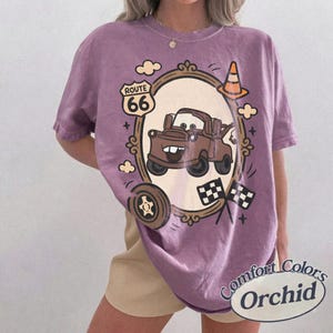 May include: Orchid Comfort Colors t-shirt featuring a cartoon image of Tow Mater from the movie Cars, inside a decorative frame. The design includes Route 66 signage, checkered flags, and a traffic cone. The shirt has a relaxed fit.