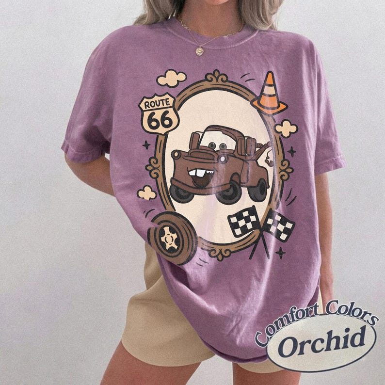 May include: Orchid Comfort Colors t-shirt featuring a cartoon image of Tow Mater from the movie Cars, inside a decorative frame. The design includes Route 66 signage, checkered flags, and a traffic cone. The shirt has a relaxed fit.