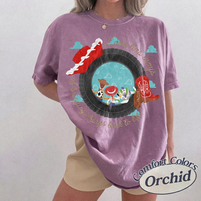 May include: A purple t-shirt featuring a graphic of a black tire framing a blue sky with clouds. Inside the tire are cartoon characters including a cowboy, a cowgirl, and a space ranger. A red hat and a red cowboy boot are also depicted.