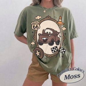 May include: Olive green t-shirt featuring a cartoon image of Mater from the movie Cars, framed by a decorative border with Route 66 signage, a traffic cone, and checkered flags. The shirt has the words "Comfort Colors Moss" printed on it.