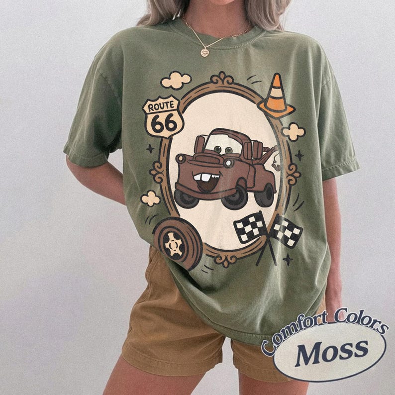 May include: Olive green t-shirt featuring a cartoon image of Mater from the movie Cars, framed by a decorative border with Route 66 signage, a traffic cone, and checkered flags. The shirt has the words "Comfort Colors Moss" printed on it.