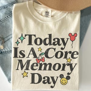 May include: Cream-coloured t-shirt with the text "Today Is A Core Memory Day" in black. The design includes stars, a cherry, a Mickey Mouse balloon, and a smiling face. The shirt is folded and rests on a pair of blue jeans.