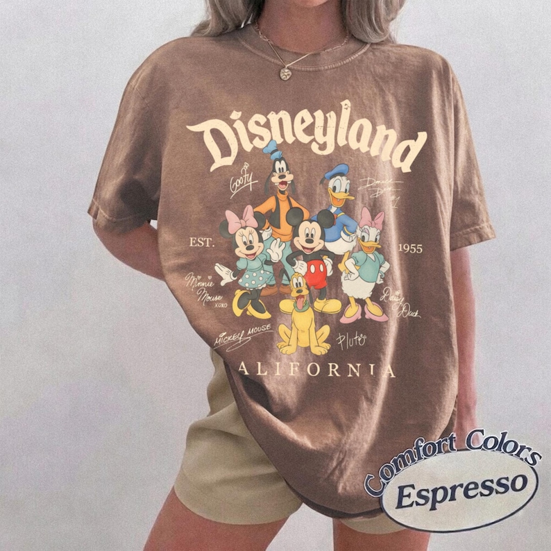 K&ouml;nnte beinhalten: Ein braunes T-Shirt mit kurzen &Auml;rmeln und einem Grafikdruck von Mickey Maus, Minnie Maus, Donald Duck, Daisy Duck, Goofy und Pluto. Der Text lautet Disneyland, Est. 1955 und California, erg&auml;nzt durch Charakter-Signaturen um die Figuren.