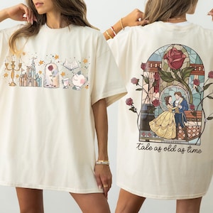 Puede incluir: Camiseta color crema con una ilustración colorida de un castillo, una rosa y otros elementos. La parte posterior de la camiseta muestra un diseño de vidriera con una pareja bailando y el texto "tale as old as time".