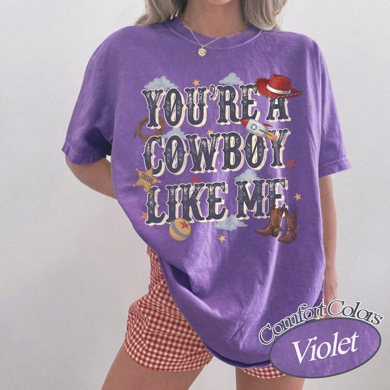 May include: A violet short-sleeved t-shirt featuring the text "YOU'RE A COWBOY LIKE ME" in a dark blue font. The graphic includes a red cowboy hat, a rocket ship, a sheriff star, a lasso, a colorful ball, and brown cowboy boots with stars.