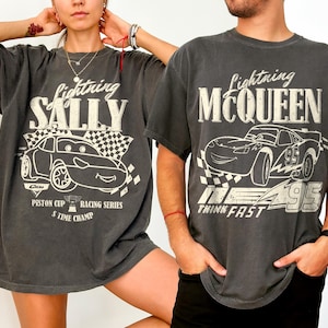 May include: Two charcoal grey t-shirts featuring Lightning McQueen and Sally Carrera from the Cars movie. Each shirt has a graphic of the character, a checkered flag, and text. The Lightning McQueen shirt says "Think Fast".