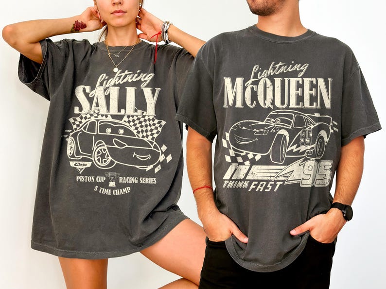 May include: Two charcoal grey t-shirts featuring Lightning McQueen and Sally Carrera from the Cars movie. Each shirt has a graphic of the character, a checkered flag, and text. The Lightning McQueen shirt says "Think Fast".