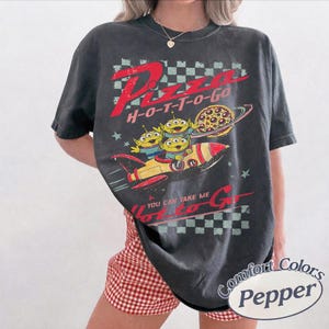 Pu&ograve; includere: T-shirt grigio scuro con grafica invecchiata. Il design include il testo "Pizza H-O-T-T-O-Go" e un cartone animato di tre alieni verdi in un razzo con un pianeta pizza. La maglietta ha un aspetto vintage.