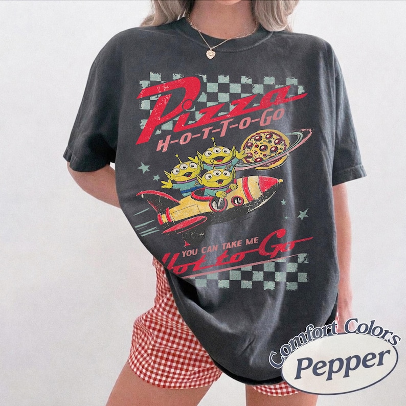 Pu&ograve; includere: T-shirt grigio scuro con grafica invecchiata. Il design include il testo "Pizza H-O-T-T-O-Go" e un cartone animato di tre alieni verdi in un razzo con un pianeta pizza. La maglietta ha un aspetto vintage.