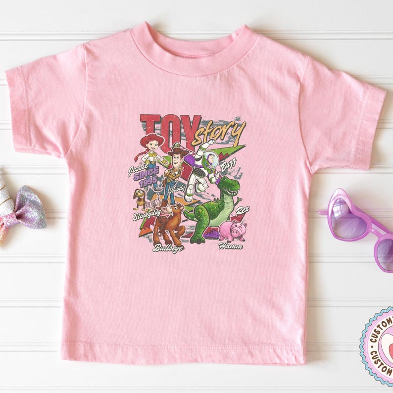 May include: A light pink short-sleeve t-shirt featuring a graphic print of Toy Story characters including Woody, Buzz Lightyear, Jessie, Rex, Hamm, Bullseye, and Slinky Dog, with the text Toy Story and character names printed in various colors.