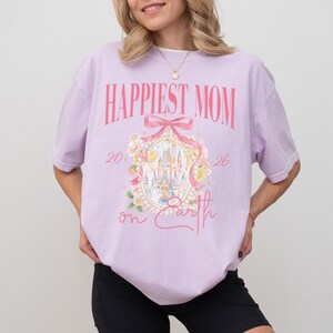 May include: A light purple t-shirt with the text "Happiest Mom" in pink. The shirt features a castle graphic with floral accents and the words "on Earth". The shirt is a casual style.