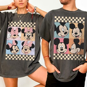 May include: Dark gray t-shirts featuring a colorful graphic of Mickey and Minnie Mouse faces in a checkerboard design. The shirts have short sleeves and a relaxed fit, suitable for casual wear.