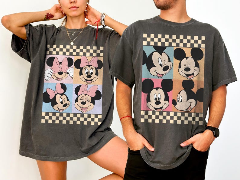 May include: Dark gray t-shirts featuring a colorful graphic of Mickey and Minnie Mouse faces in a checkerboard design. The shirts have short sleeves and a relaxed fit, suitable for casual wear.
