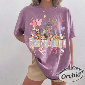 May include: Orchid-colored t-shirt featuring a Disneyland graphic with Mickey Mouse, Minnie Mouse, Donald Duck, and other Disney characters. The shirt has the words "Disneyland" and "Comfort Colors Orchid" printed on it.