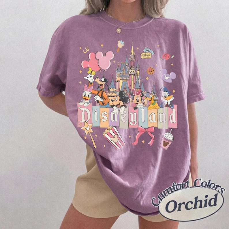 May include: Orchid-colored t-shirt featuring a Disneyland graphic with Mickey Mouse, Minnie Mouse, Donald Duck, and other Disney characters. The shirt has the words "Disneyland" and "Comfort Colors Orchid" printed on it.