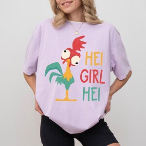 Peut inclure: T-shirt lilas avec un dessin de coq de dessin anim&eacute; aux accents rouges, verts, jaunes et oranges. Le texte "HEI GIRL HEI" est imprim&eacute; en jaune, rouge et vert. Le t-shirt est ample.