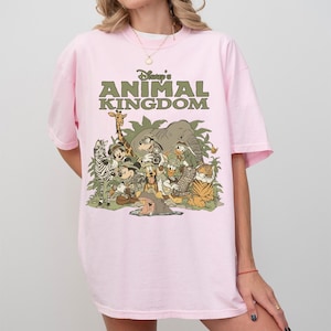 Puede incluir: Camiseta rosa claro con el texto "Disney's Animal Kingdom" en verde. El gr&aacute;fico presenta personajes y animales de Disney, incluyendo una cebra, una jirafa, un elefante y un tigre.
