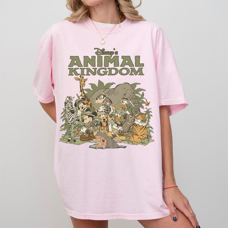 Puede incluir: Camiseta rosa claro con el texto "Disney's Animal Kingdom" en verde. El gr&aacute;fico presenta personajes y animales de Disney, incluyendo una cebra, una jirafa, un elefante y un tigre.