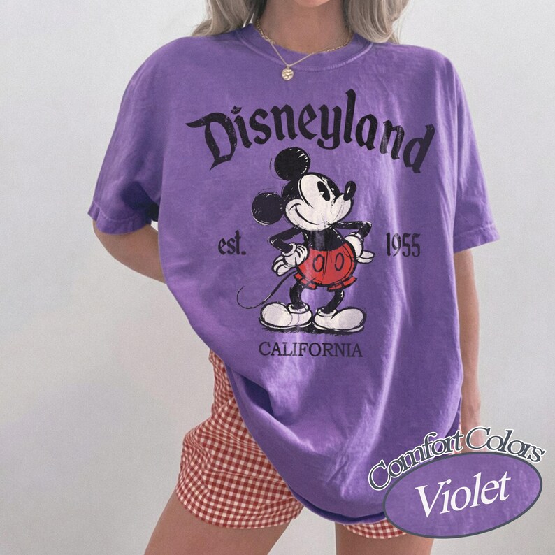 Może przedstawiać: Fioletowa koszulka Comfort Colors z grafiką Myszki Miki i napisem "Disneyland". Koszulka zawiera r&oacute;wnież napis "est. 1955 CALIFORNIA".
