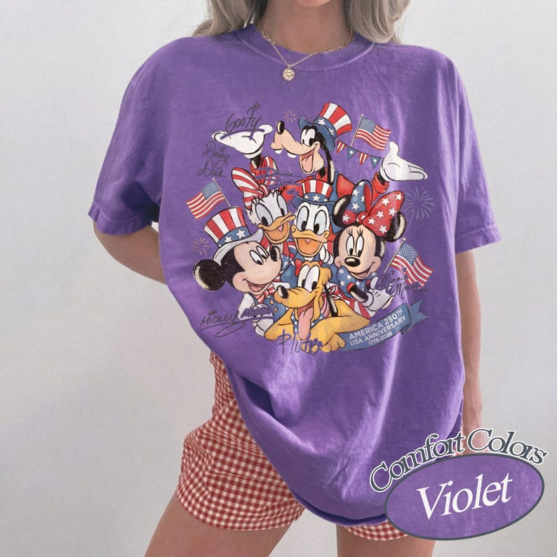 Puede incluir: Camiseta violeta Comfort Colors con un gr&aacute;fico de los personajes de Disney Mickey Mouse, Minnie Mouse, Donald Duck, Daisy Duck, Goofy y Pluto, celebrando el 250 aniversario de EE. UU. El dise&ntilde;o incluye banderas americanas y sombreros patri&oacute;ticos.