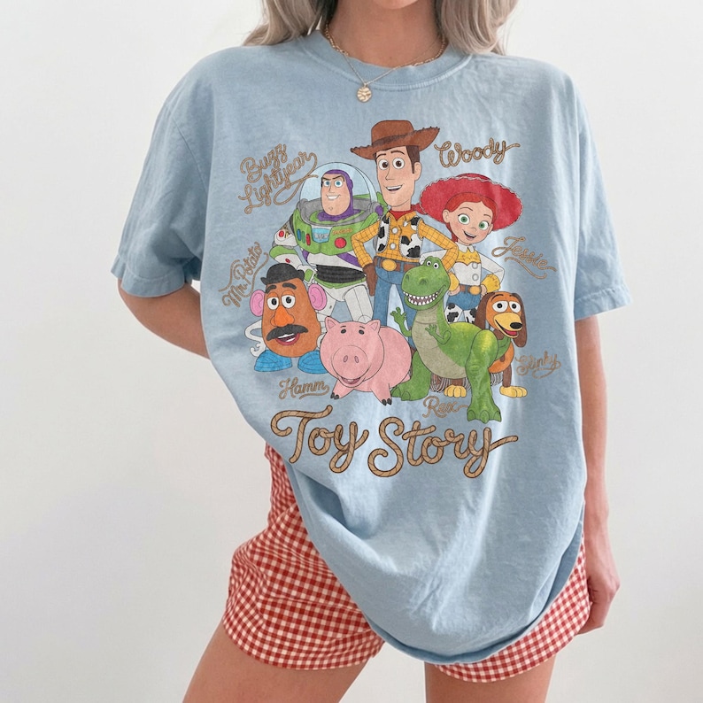 May include: A light blue t-shirt featuring a graphic print of Toy Story characters including Woody, Buzz Lightyear, Jessie, Mr. Potato Head, Hamm, Rex, and Slinky Dog. The text Toy Story is printed in a brown cursive font below the characters.