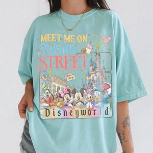 May include: Light blue t-shirt with a colorful graphic featuring Disney characters and the text "Meet Me On Main Street Disneyworld." The design includes Mickey Mouse, Minnie Mouse, Donald Duck, and a castle. The shirt has a relaxed fit.