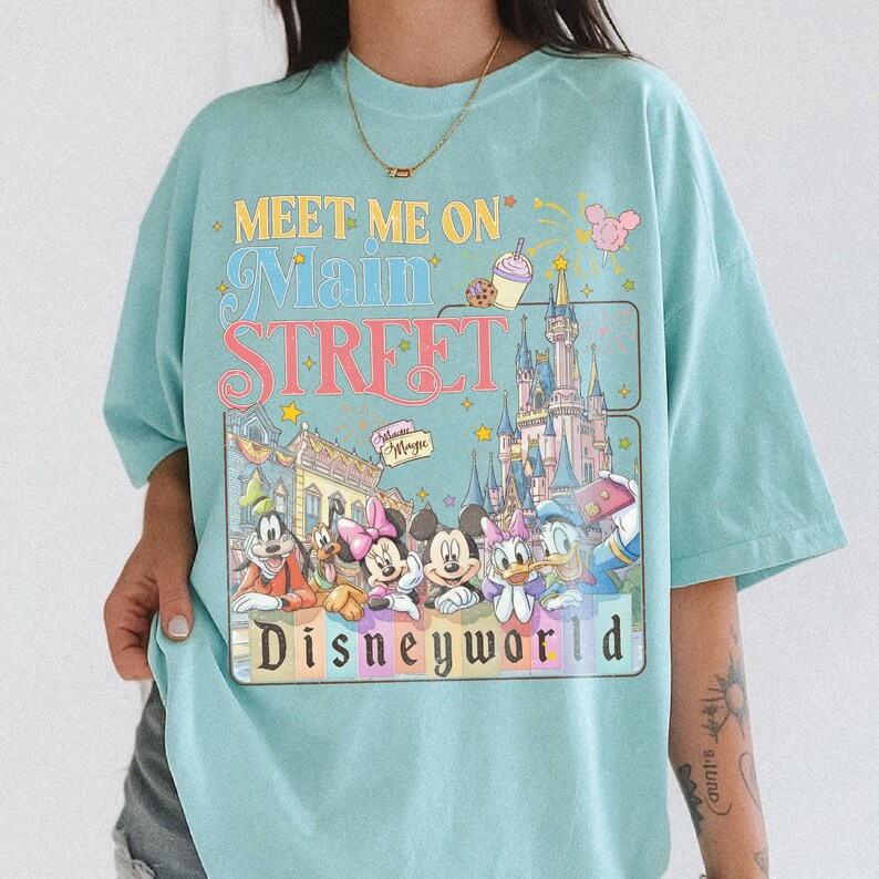 May include: Light blue t-shirt with a colorful graphic featuring Disney characters and the text "Meet Me On Main Street Disneyworld." The design includes Mickey Mouse, Minnie Mouse, Donald Duck, and a castle. The shirt has a relaxed fit.