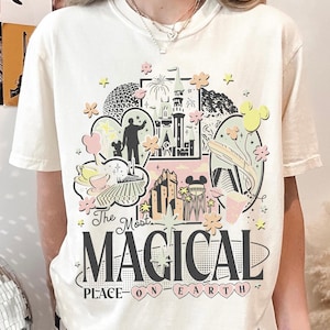 May include: Off-white t-shirt with a vibrant graphic design that reads "The Most Magical Place on Earth." The design incorporates illustrations of a castle, fireworks, and other iconic imagery. The shirt has a crew neck and short sleeves.