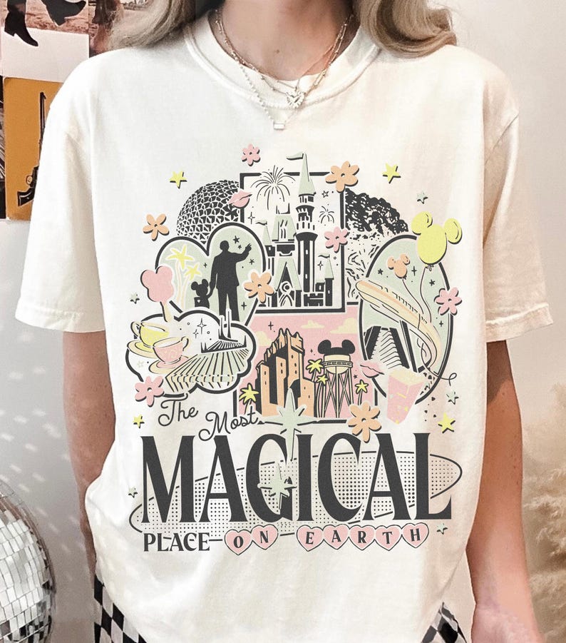 May include: Off-white t-shirt with a vibrant graphic design that reads "The Most Magical Place on Earth." The design incorporates illustrations of a castle, fireworks, and other iconic imagery. The shirt has a crew neck and short sleeves.