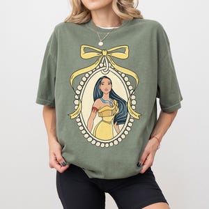 May include: Olive green t-shirt featuring a framed illustration of a woman with long black hair and a yellow dress. The illustration is surrounded by a yellow bow and a pearl-like border. The shirt has short sleeves.