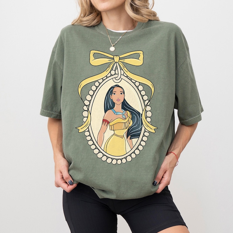May include: Olive green t-shirt featuring a framed illustration of a woman with long black hair and a yellow dress. The illustration is surrounded by a yellow bow and a pearl-like border. The shirt has short sleeves.