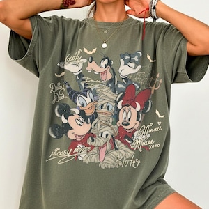 May include: Khaki green t-shirt with a graphic of Disney characters, including Mickey Mouse, Minnie Mouse, Goofy, and Daisy Duck, in Halloween costumes. The shirt has the names of the characters written in script.