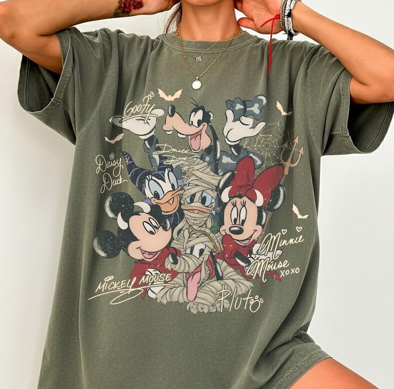 May include: Khaki green t-shirt with a graphic of Disney characters, including Mickey Mouse, Minnie Mouse, Goofy, and Daisy Duck, in Halloween costumes. The shirt has the names of the characters written in script.