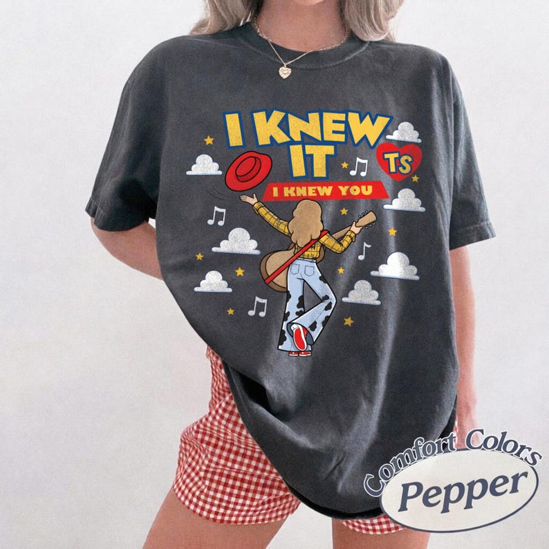 May include: A charcoal grey t-shirt featuring a graphic of a person with long hair playing a guitar, wearing a yellow plaid shirt and cow-print trousers. The text reads I KNEW IT and I KNEW YOU, accompanied by a red hat, a red heart, and white clouds.